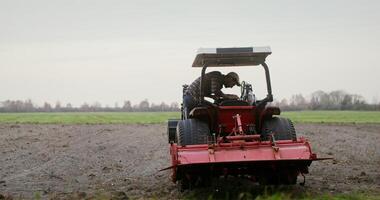 A young black haired man with a beard gets behind the wheel of a small agricultural tractor, lowers the plow and starts plowing the field. filming from behind photo