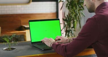 A young dark haired man with stubble on his face is typing on a laptop with a green screen while sitting at a table in a coffee shop. Chroma key, copy space. Videography from the side photo