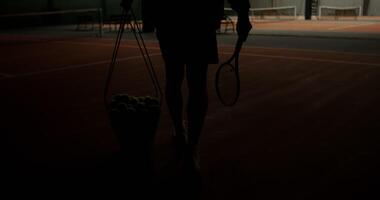 The tennis court lights come on and a man comes in, getting ready for the game, carrying a basket full of tennis balls. Videography of male legs with a basket from behind, close up photo