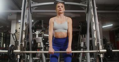 Young beautiful woman with athletic physique, dressed in a sports uniform, doing bends with a barbell on the exercise machine, doing in the gym. Front videography, close up photo