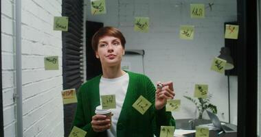 A young woman of European appearance is developing a strategy for a startup, drawing on a glass board with a marker and sticking stickers on it, working in a modern office, close up photo