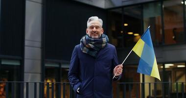 Respectable grey haired man smiling looking at the camera, holding a yellow blue flag of Ukraine, standing near an office building on a cold evening. Chroma key, mockup. in 4k, red komodo photo