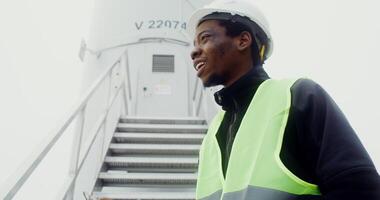 African American man in a green vest and hard hat takes notes in a glider while standing near the entrance to an industrial wind turbine, close up, dynamic . in 4k, red komodo photo