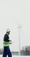 A young African American man in a work uniform with a helmet on his head is diagnosing a wind turbine using a digital tablet, vertical . in 4k, red komodo photo