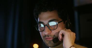 Young curly haired brunette man, wearing glasses, talking on a microphone with headphones and looking at a laptop in the dark, close up. Customer support photo