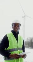 A young African American man in work uniform with hard hat on his head smiles while looking at camera while standing near a wind turbine on a winter snowy day, vertical view. in 4k, red komodo photo
