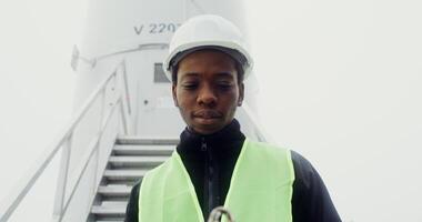 A young African American man in a green vest and hard hat smiles while looking at the camera, standing near the entrance to an industrial wind turbine. in 4k, red komodo photo