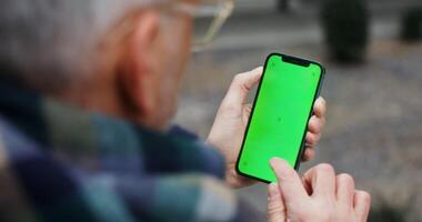 A gray haired man in a voluminous scarf uses a mobile phone with a green screen, an unrecognizable person, a top view from behind the man's shoulder. Chroma key, mockup. in 4k, red komodo photo