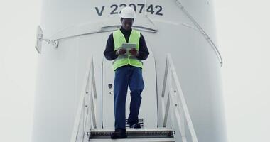A young African American man in work gear uses a tablet with a serious look as he walks down the steps of a wind turbine during an inspection. in 4k, red komodo photo