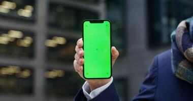 A warmly dressed man holds a green screen phone as he stands outside an office building. Copy space, mockup, close up of the phone, no face. in 4k, red komodo photo