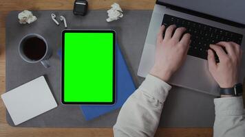 A digital tablet with a green screen lies on the desktop next to an open laptop, on the keyboard of which a man is typing, close up, top view, no face. Mockup, chroma key, copy space photo