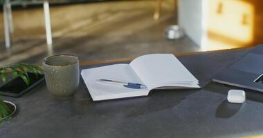 An open notebook with a pen lie on the table next to a laptop, a mug and a case for wireless headphones, close up, no people. Workplace for remote work photo
