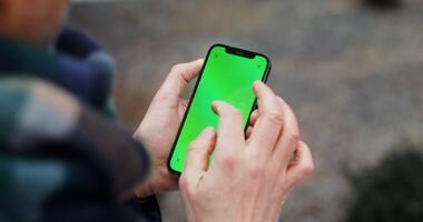 A warmly dressed man uses a phone with a green screen, swiping his finger across the screen, close up, no face, view from behind the man. Copy space, mockup. in 4k, red komodo photo