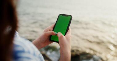 A woman is typing on smartphone with a green screen while sitting on the rocks on the seashore. Close up phone in the woman's hands, no face. Chroma key, mockup, copy space. 4k , red komodo photo