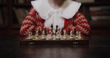 A young woman of European appearance plays a chess game alone, thinking long concentratedly between moves, sitting at a table in the library, close up photo