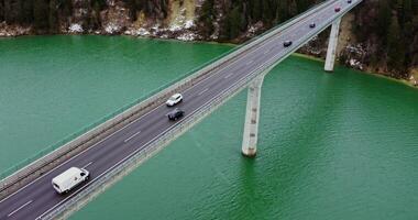 Top view of a road bridge with a two lane asphalt road with cars passing on it. drone footage of a long bridge across an emerald green river to hills covered in snowy forest. photo