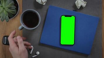 A man takes a cup of hot coffee, standing on desktop next to a mobile phone with a green screen, lying on a notebook with pieces of paper crumpled next to it, close up. Mockup, chroma key, copy space photo
