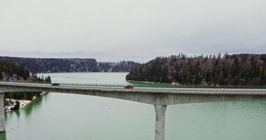 View of wide river flowing among spruce forests and fields covered with snow, and a narrow concrete road bridge connecting the banks, filming from a bird's eye view, from a drone photo