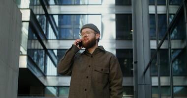 A young stylishly dressed man in a knitted hat is talking on a mobile phone while walking near high rise office buildings with large stained glass windows. in 4k, red komodo photo
