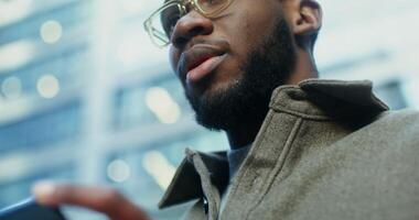 An African American man uses a mobile phone while standing near modern buildings. Close up of a focused male face, viewed from below. in 4k, red komodo photo