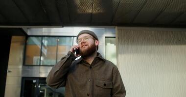 A young man with a beard, dressed in casual clothes and a knitted hat, is talking on a mobile phone while walking in the courtyard of a modern glass building, close up. in 4k, red komodo photo
