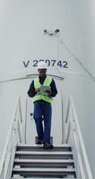 A young African American man wearing a white hard hat and green vest uses a tablet while walking down the steps of a wind turbine during an inspection. in 4k, red komodo photo