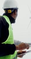 African American man in a work uniform with a hard hat on his head uses a tablet while standing near a wind turbine and looking at it, winter day, close up, vertical . in 4k, red komodo photo