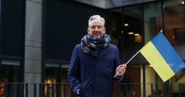 A respectable gray haired warmly dressed man smiling looking at the camera, holding a yellow blue flag of Ukraine, standing near an office building. Chroma key, mockup. in 4k, red komodo photo