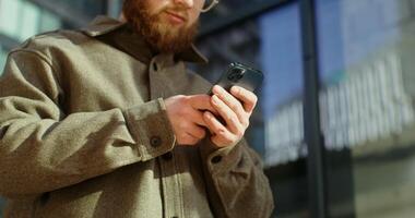 A young man in a knitted hat with a voluminous red beard is typing text on a mobile phone, standing near a glass building on a sunny day, close up. in 4k, red komodo photo