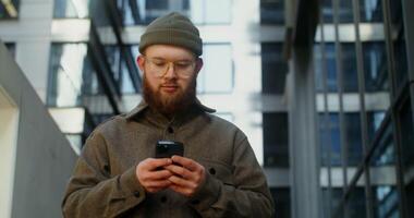 A young man in a knitted hat with a voluminous red beard is typing on a mobile phone while walking around the business center of the city, close up. in 4k, red komodo photo