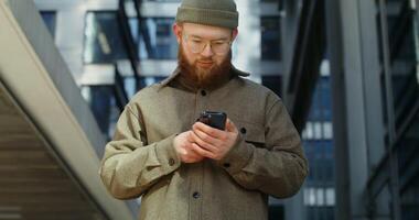 A bearded stylishly dressed man in glasses uses a mobile phone with a focused look, standing in the business center of the city, close up. in 4k, red komodo photo