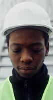 A young African American man in a white hard hat and a green vest looks at the camera with a smile, raising his head, close up. in 4k, red komodo photo