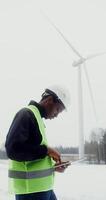 A young African American man in a work uniform with a hard hat on his head is diagnosing a wind turbine using a digital tablet on a winter day, close up, vertical . in 4k, red komodo photo