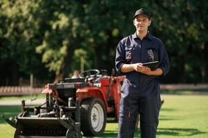 In ,,cap, holding digital tablet., Man is with utility tractor with grass cutter and aerator equipment on the field photo