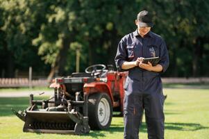 In ,cap, holding ,digital,, tablet. Man is with utility tractor with grass cutter and aerator equipment on the field photo