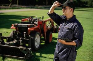 Black digital tablet in hands.,,, Man is ,with utility tractor with grass cutter and aerator equipment on the field photo