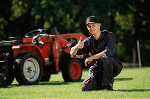 Sitting, with thumb up. Man is, with ,utility tractor with grass cutter and aerator equipment on the field photo
