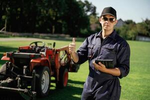 Shows ,,thumb, up. Man is with utility tractor with grass cutter and aerator equipment on the field photo