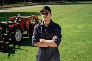Standing with ,arms crossed. Man is with utility tractor with grass cutter and aerator equipment on the field photo