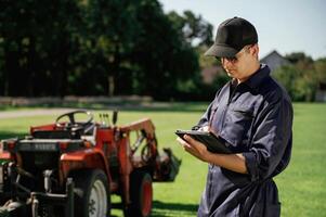 Black digital tablet in ,hands. Man is with utility tractor with grass cutter and aerator equipment on the field photo