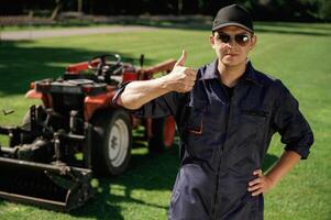Shows thumb up. Man, is with utility tractor with grass cutter and aerator equipment on the field photo