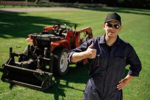 Shows, thumb up. Man is with utility tractor with grass cutter and aerator equipment on the field photo