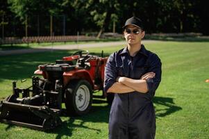Standing ,with arms crossed. Man is with utility tractor with grass cutter and aerator equipment on the field photo