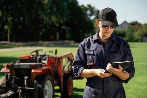 Black ,digital tablet in hands. Man is with utility tractor with grass cutter and aerator equipment on the field photo