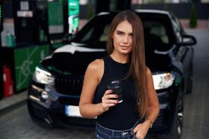 Black, cup of the drink, in, hand. A young woman at a gas station with her car photo