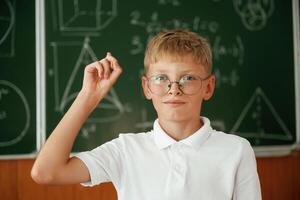 Kid in glasses ,,,is standing in ,the c,lassroom against the blackboard with graphs, hand up photo
