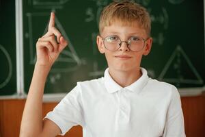 Kid in glasses, is, standing in the classroom against the blackboard with graphs, hand up photo