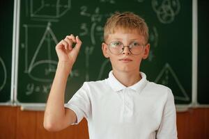 Kid ,in glasses is standing in the classroom against the blackboard with graphs, hand up photo