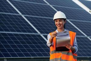 Checking the ,,,data, with notepad. Female worker engineer in uniform is near solar panels photo