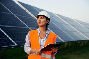 Checking, the data, with notepad. Female worker engineer in uniform is near solar panels photo